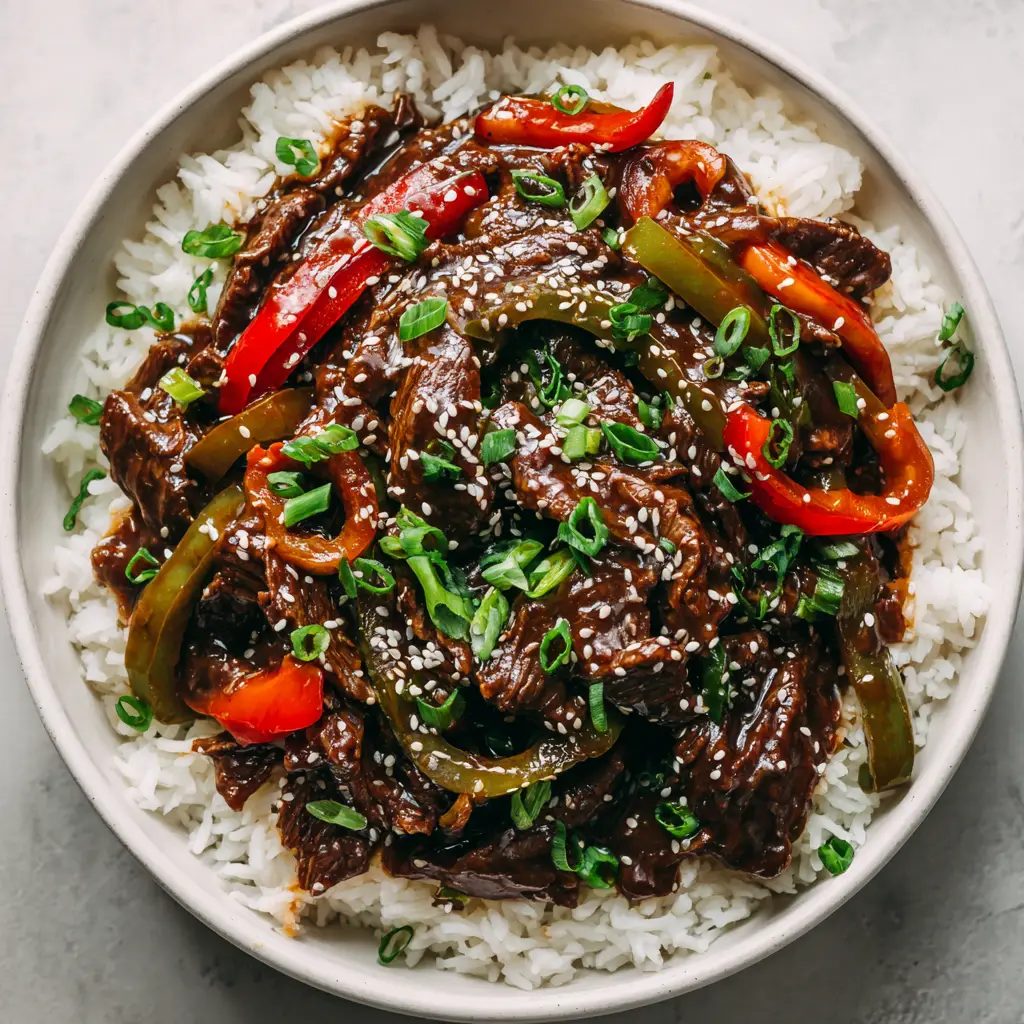 Slow cooker pepper steak garnished with toasted white sesame seeds and raw green scallions, plated in a shallow white ceramic bowl.