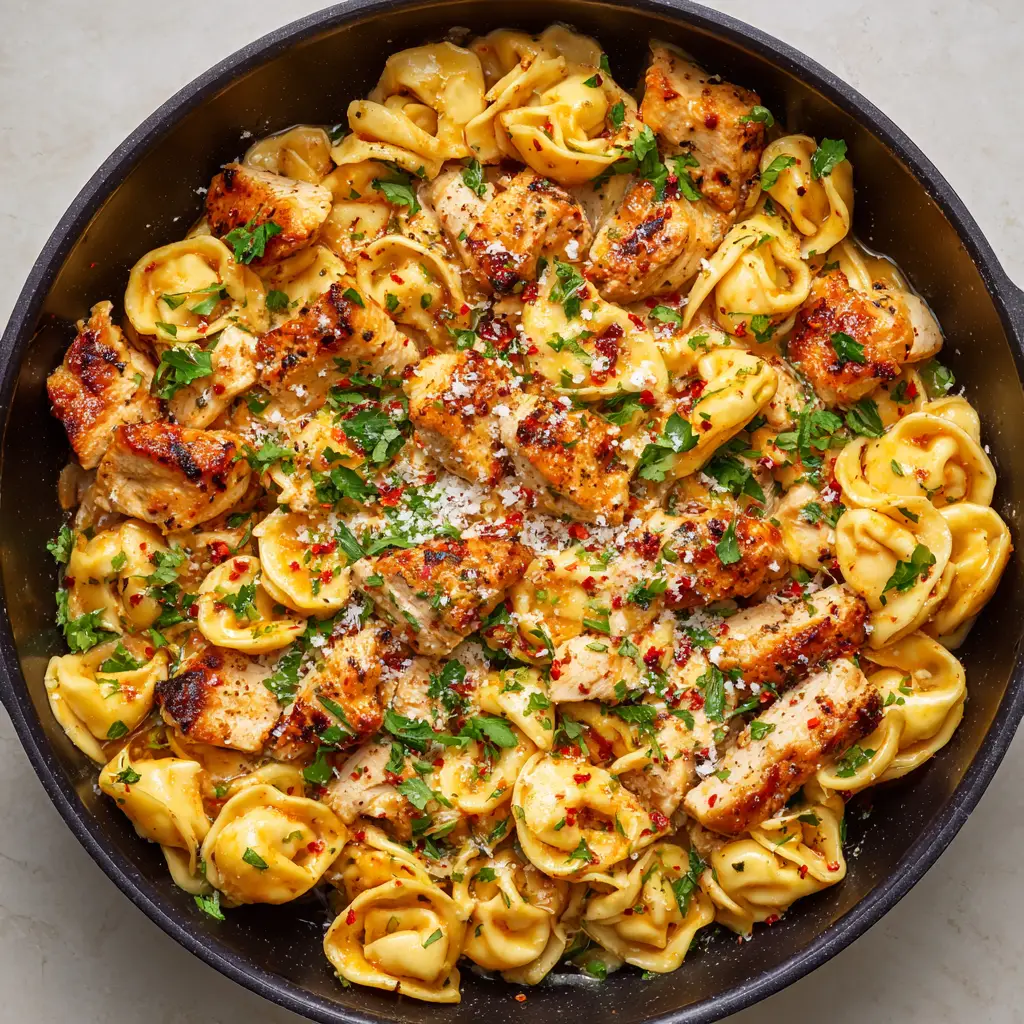 A cast-iron skillet filled with cheese tortellini, chicken, bright red pepper flakes, fine white Parmesan cheese, and vibrant green parsley.